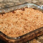 A warm golden-brown apple crisp topped with a crunchy oat crumble, servied in a rustic baking dish. The filling glistens with baked apples and a hint of cinnamon